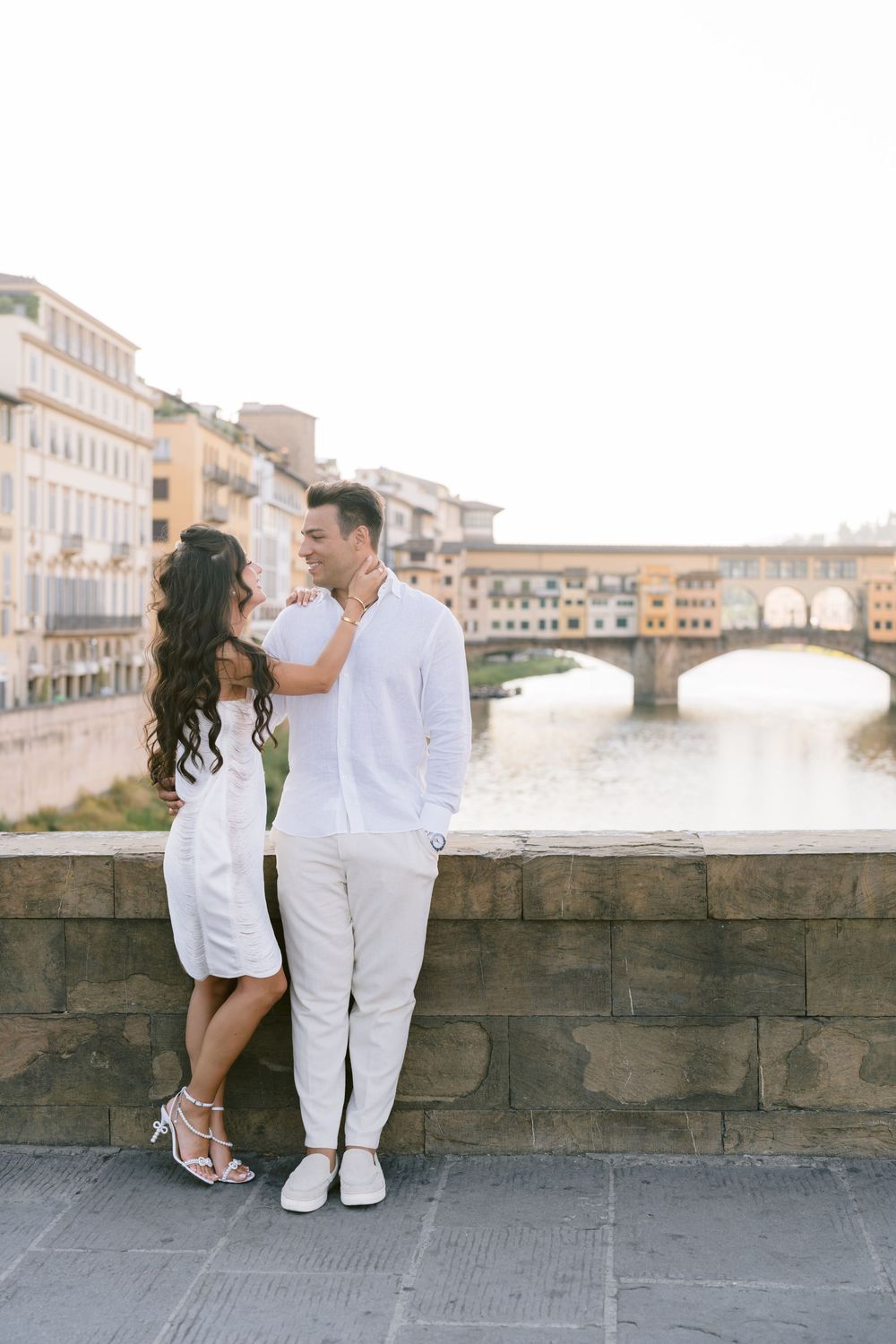 Couple in white outfits posing by the Arno River with Ponte Vecchio bridge in Florence, Italy.