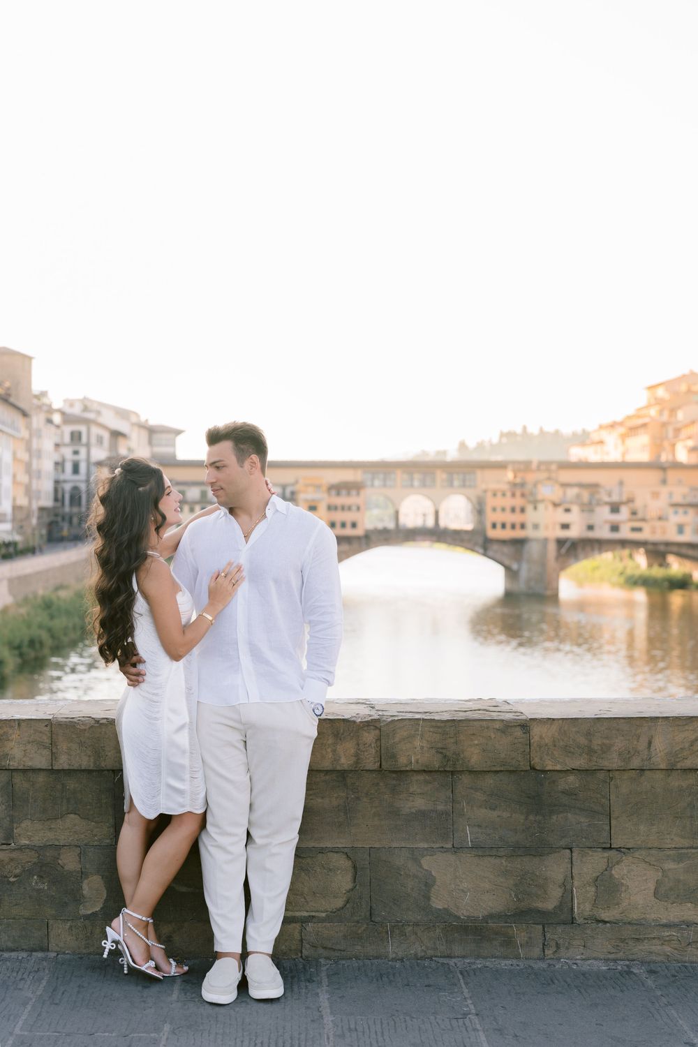 Couple in white outfits embracing by the Ponte Vecchio bridge over the Arno River in Florence, Italy.