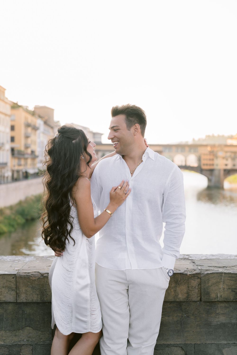 Couple in white outfits embracing on a bridge over the Arno River with Ponte Vecchio in Florence, Italy.