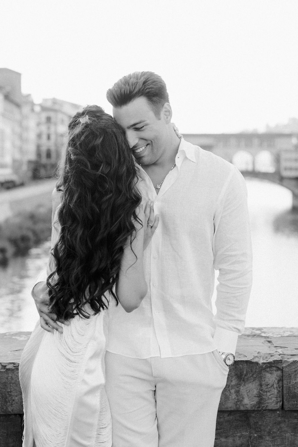 Black and white photo of a smiling couple embracing on a bridge in Florence, Italy, with the Arno River in the background.
