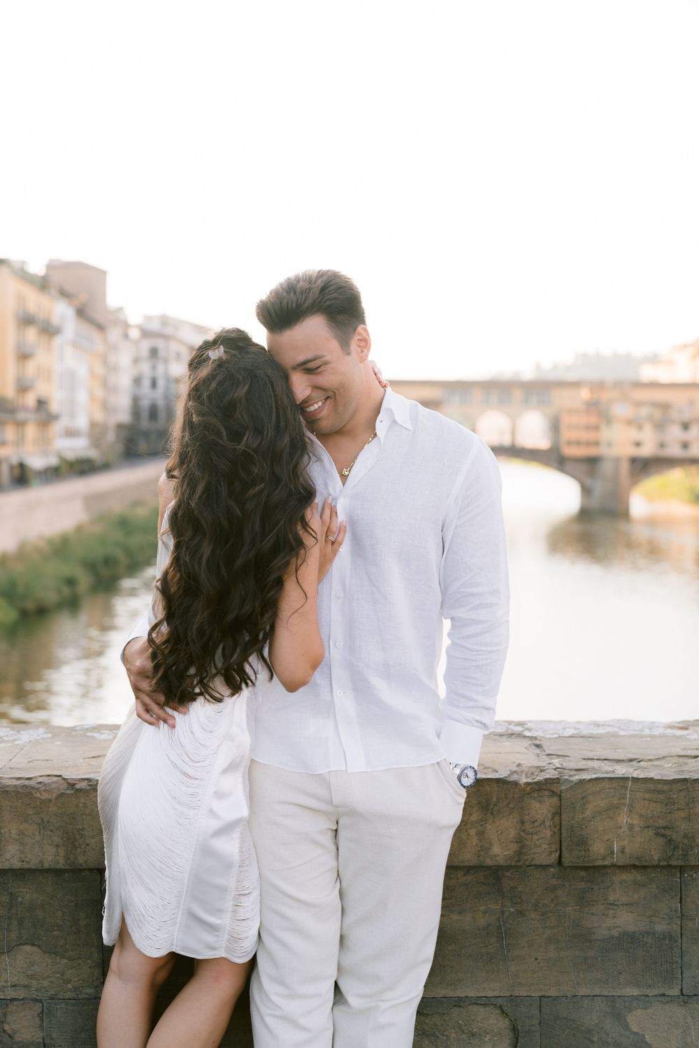 Romantic couple in white outfits embracing on a bridge over the Arno River in Florence, Italy.
