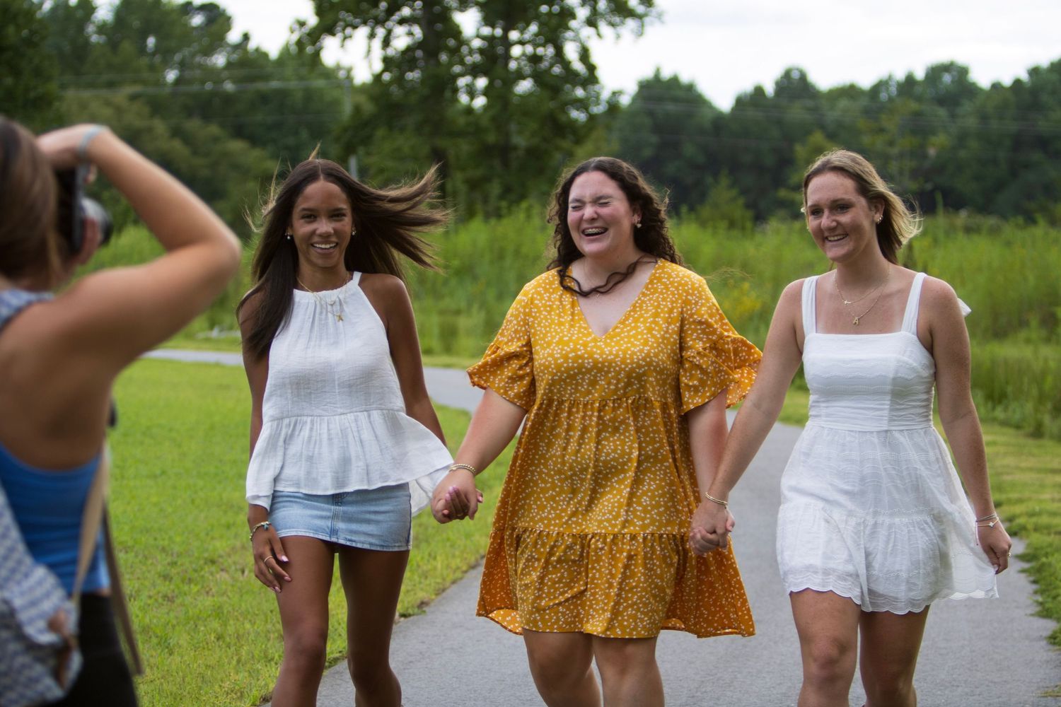 Three women holding hands and laughing while walking outdoors as a photographer captures their joyful moment.