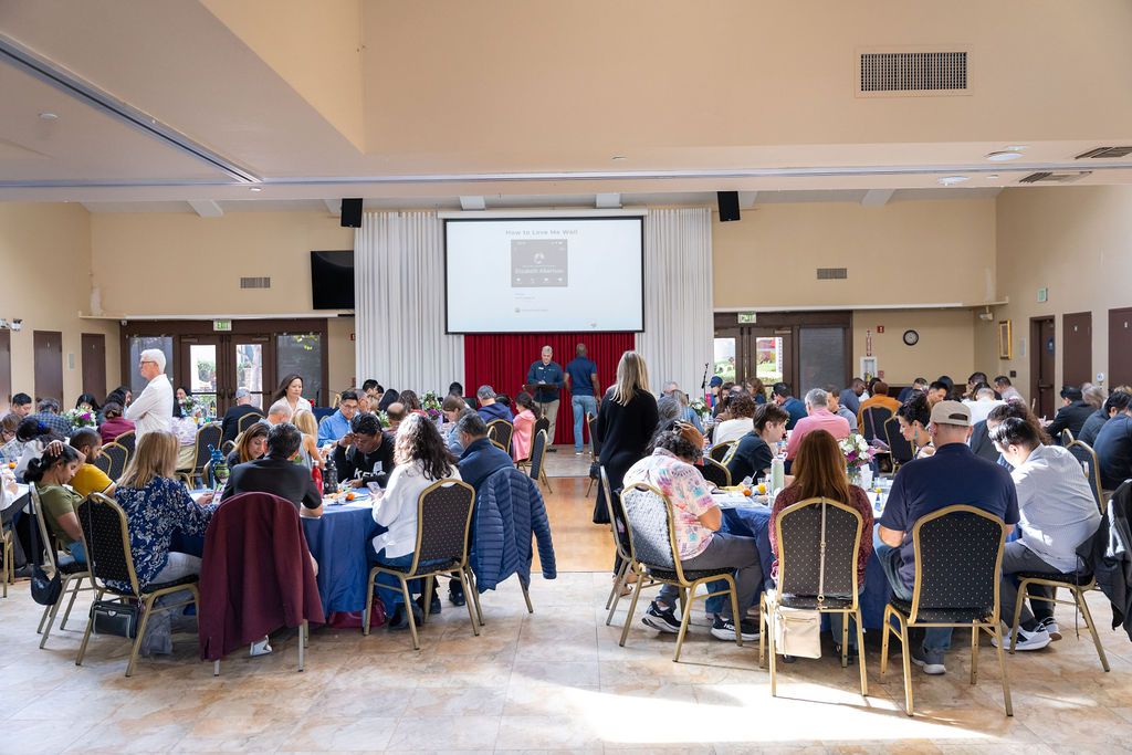 Large conference hall filled with attendees seated at round tables during a formal event with a presenter on stage.