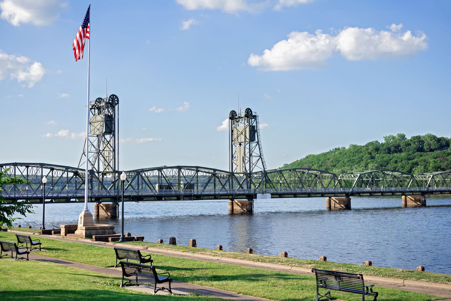 Historic lift bridge spanning a wide river with an American flag and park benches in the foreground.