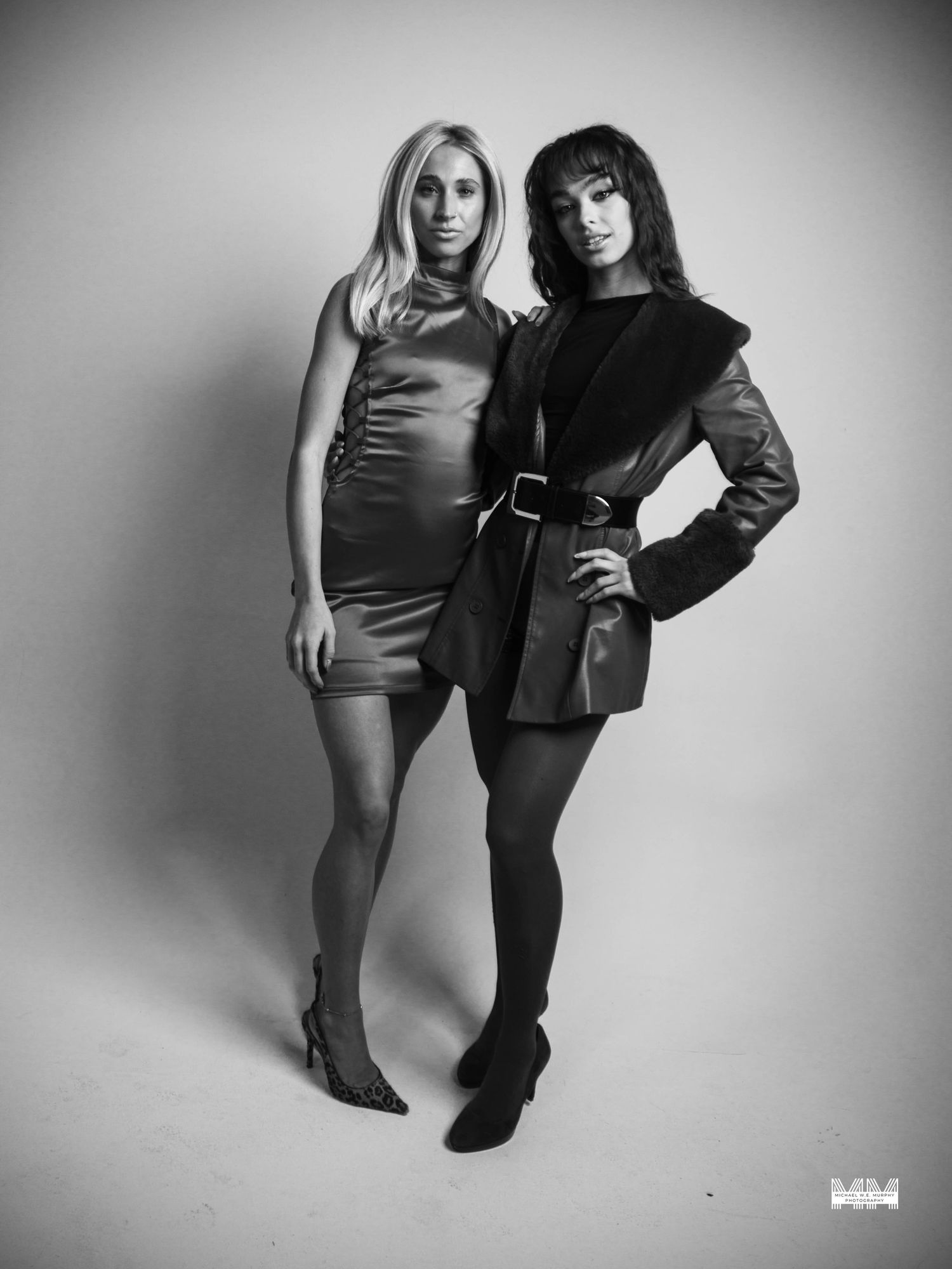 Two women pose in a studio, one in a mini dress and heels, the other in a belted leather jacket and boots.