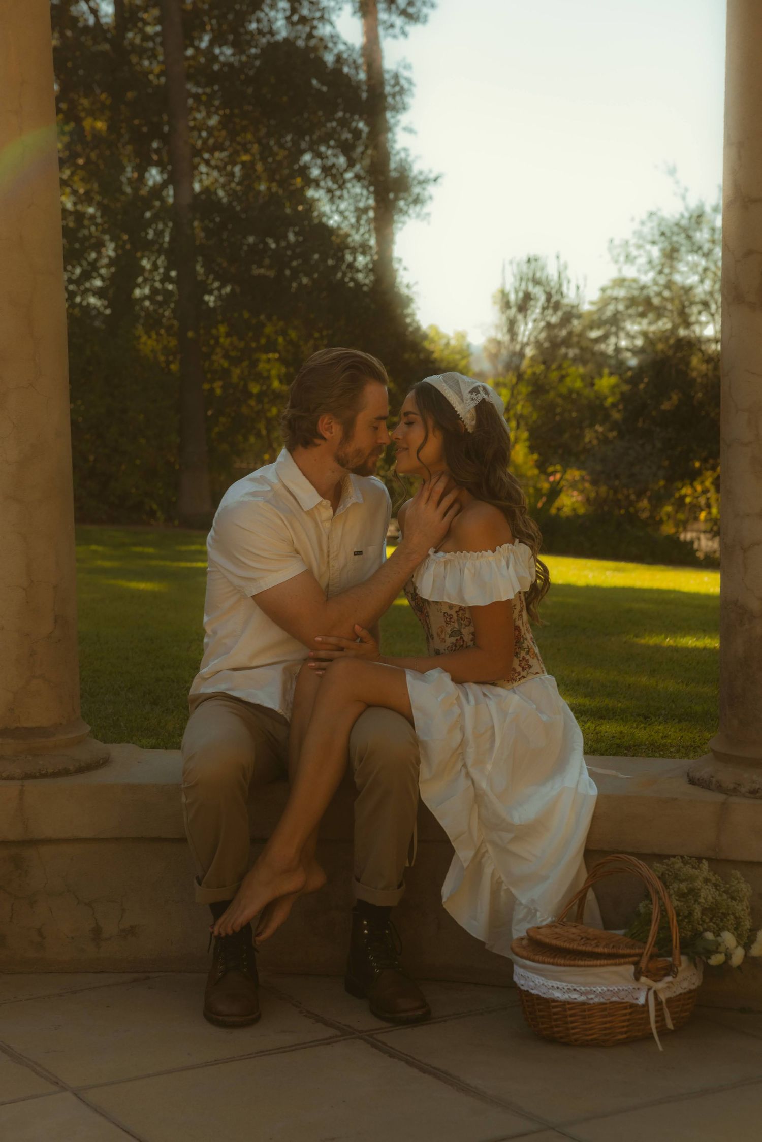Romantic couple sharing an intimate moment at golden hour, sitting between stone columns in a lush garden with a wicker basket nearby.