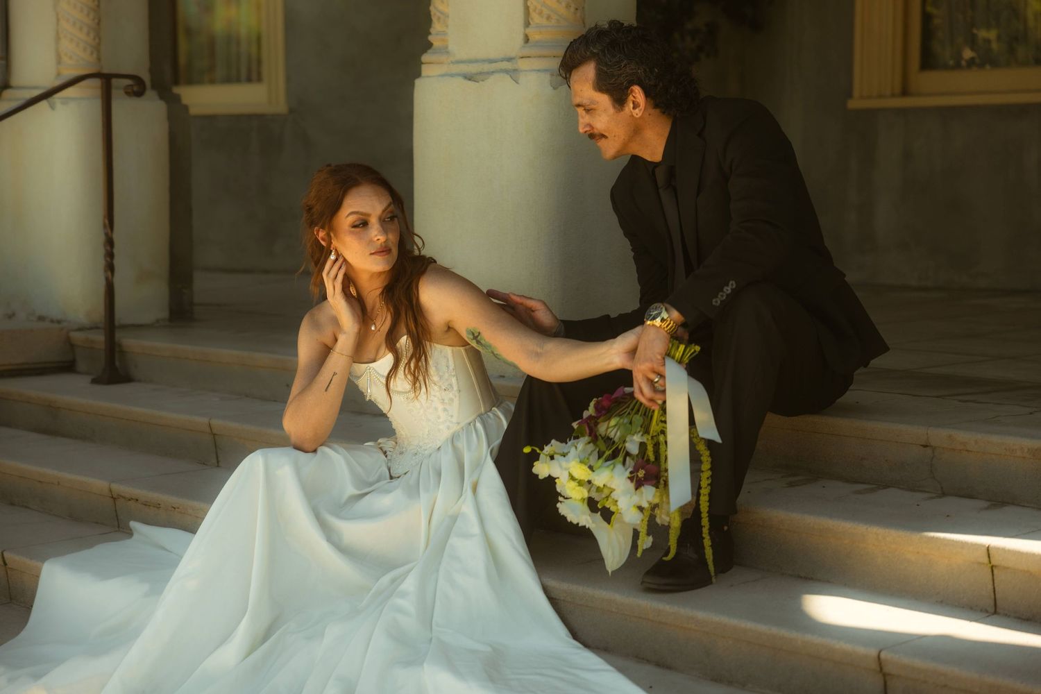 A bride in a white gown sits on stone steps as a groom in a black suit kneels beside her holding a bouquet.
