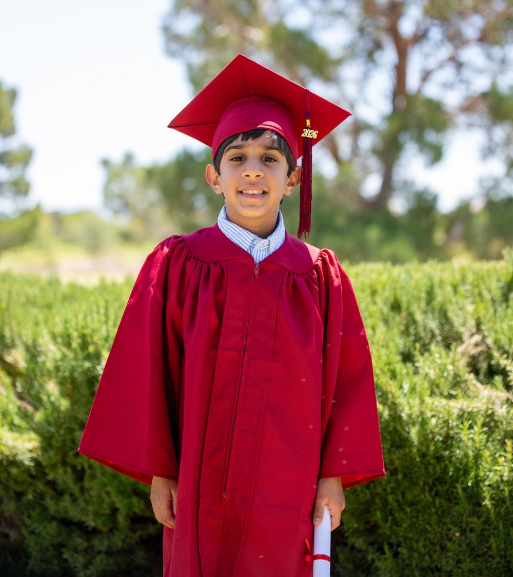 Young boy in red graduation cap and gown holding diploma, smiling outdoors with green trees in background.