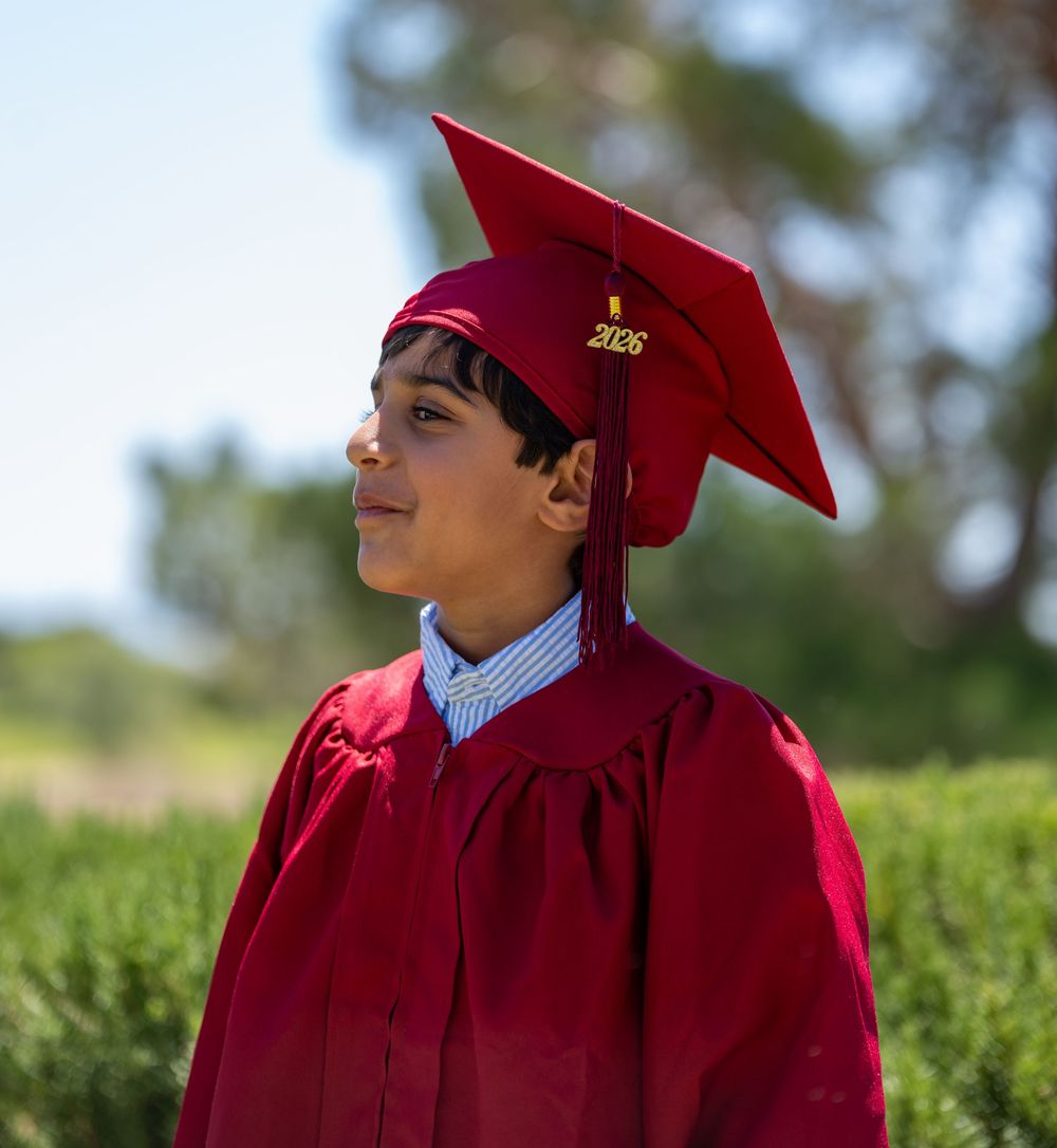 Young boy in red graduation cap and gown smiling outdoors with green trees in background.