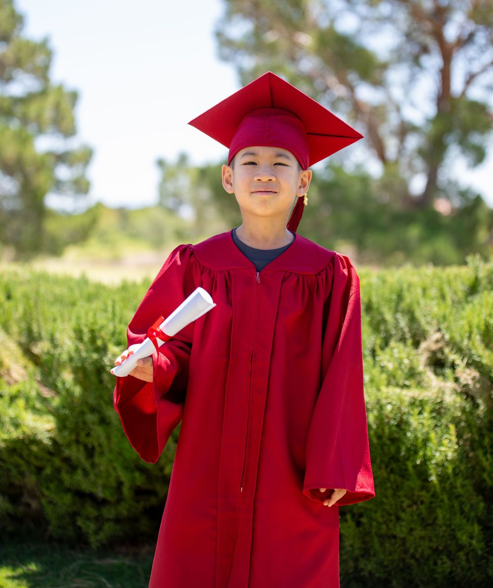 Young boy in red graduation cap and gown holding diploma outdoors.