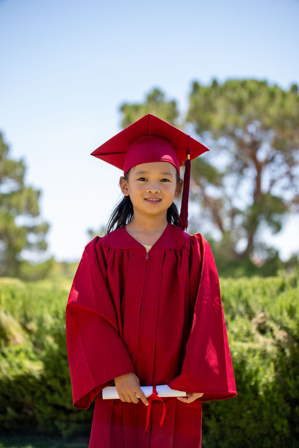 Young girl in red graduation cap and gown holding diploma outdoors.