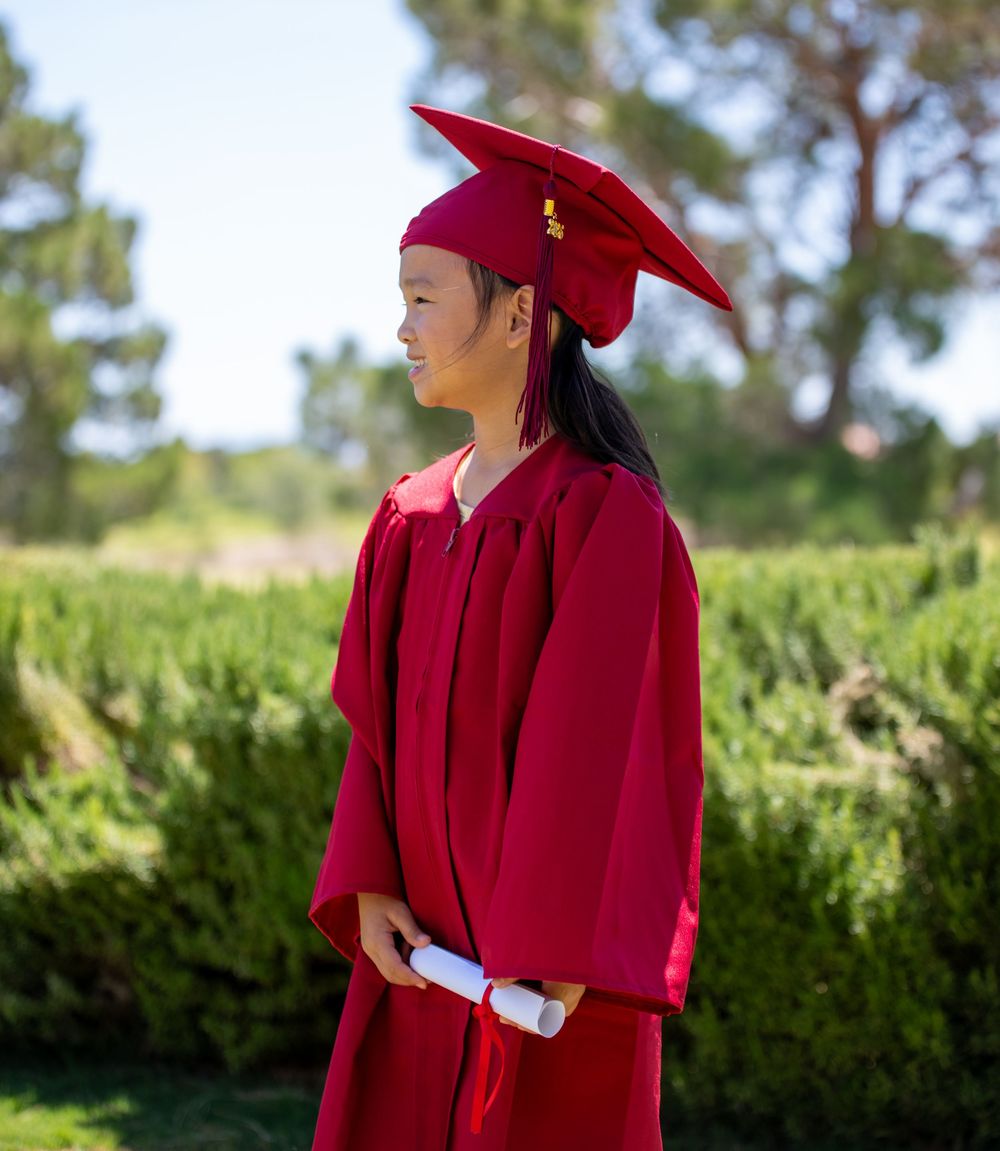 Young girl in red graduation cap and gown holding diploma outdoors.