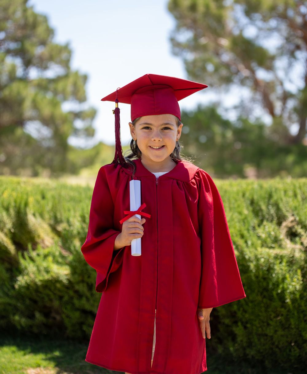 Smiling child in red cap and gown holding diploma scroll outdoors.