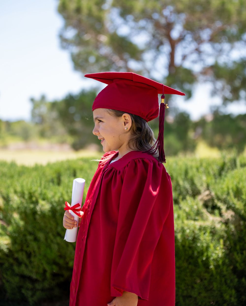Young girl in red graduation cap and gown holds diploma outdoors, smiling proudly at kindergarten ceremony.