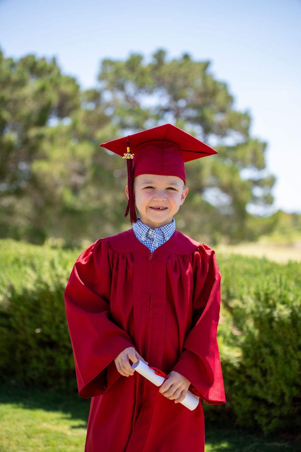 Young boy in red graduation cap and gown holding diploma scroll outdoors on a sunny day.