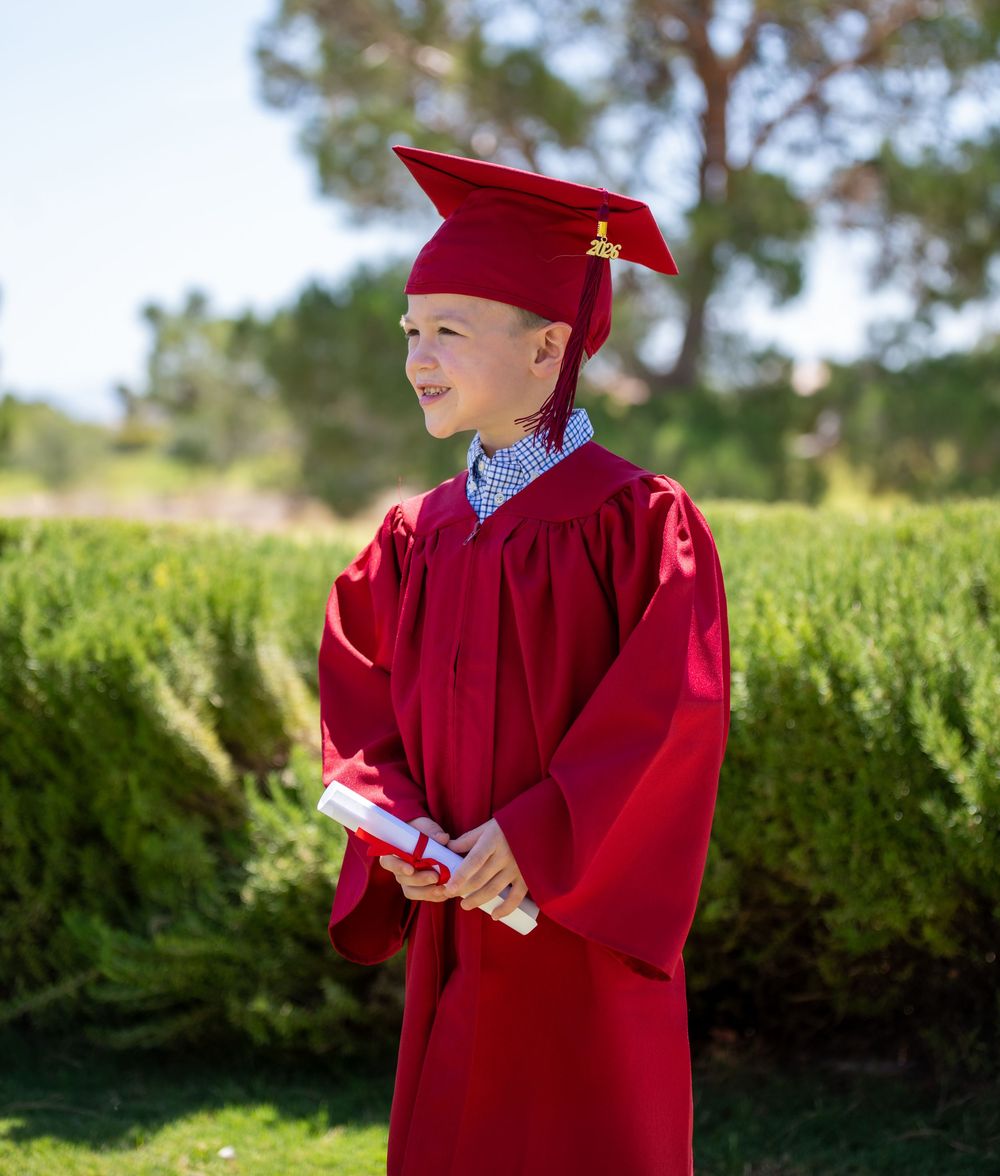 Smiling boy in red graduation cap and gown holding diploma outdoors.