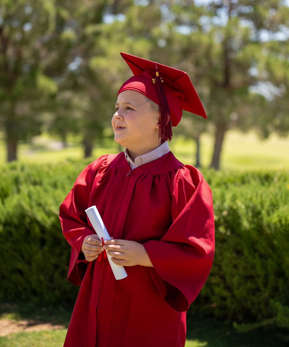Young child in red graduation cap and gown holding diploma outdoors among green trees and shrubs.