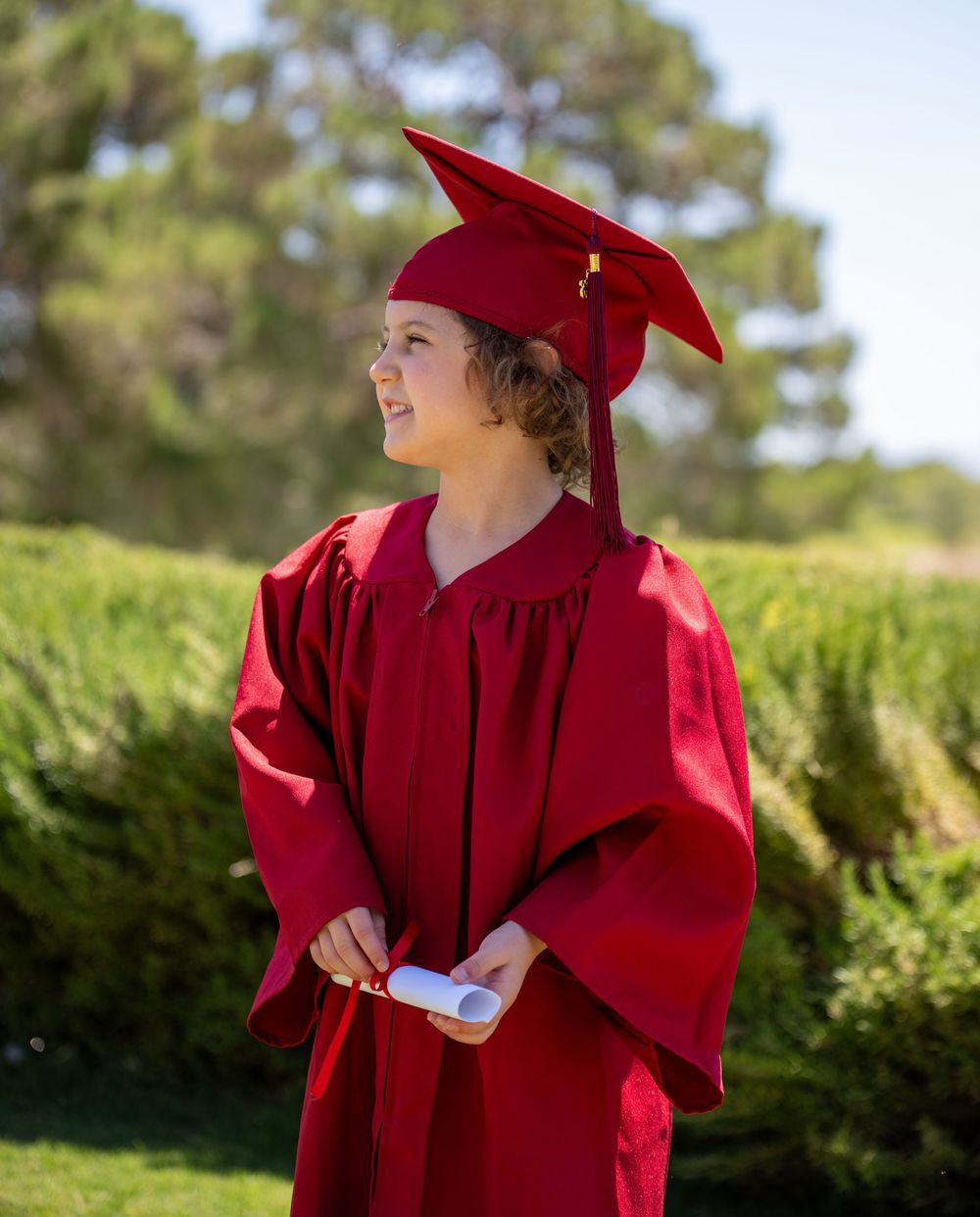Child in red graduation cap and gown holding diploma while looking up outdoors with green trees behind.