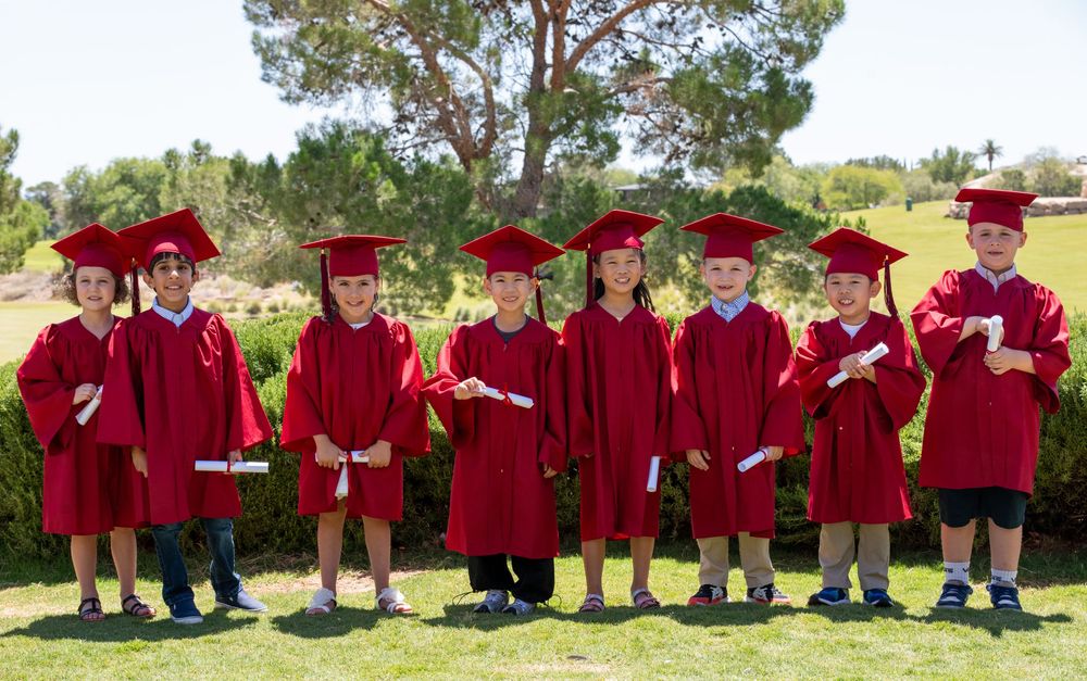 Seven young children in red graduation caps and gowns holding diplomas pose outdoors for a preschool graduation photo.