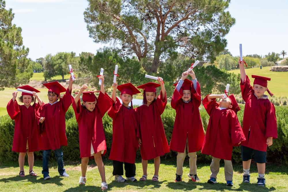 Young children in red graduation gowns and caps celebrate outdoors, holding diplomas aloft in a joyful kindergarten graduation ceremony.