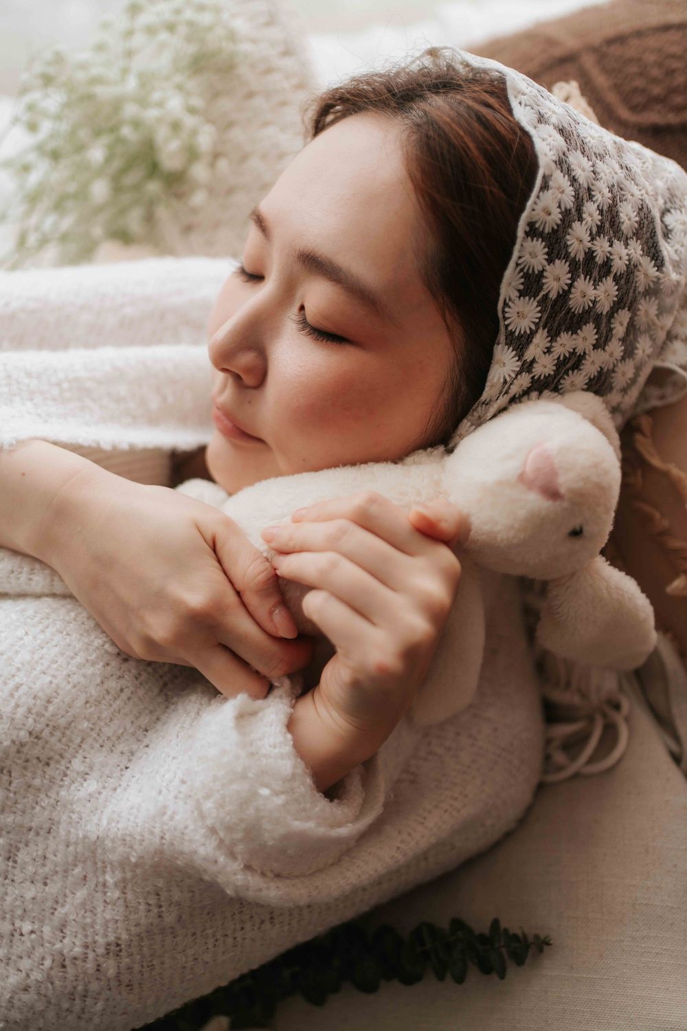 A young Asian woman rests peacefully with eyes closed, hugging a small white stuffed animal toy while wrapped in a cozy blanket.