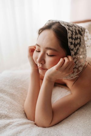 A young Asian woman with closed eyes rests peacefully on a bed, wearing a patterned headscarf, hands gently cradling her face.