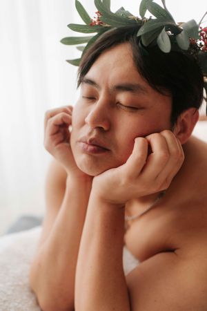 Shirtless person with closed eyes and floral crown of green leaves and red berries resting face in hands.