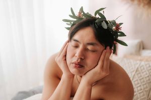 Person wearing a floral crown with green leaves and red berries, resting face in hands with eyes closed.