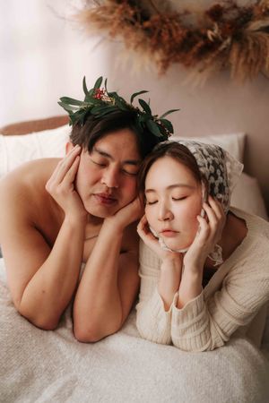 A couple with closed eyes resting together, man wearing a floral crown, woman in a knit sweater and headscarf.