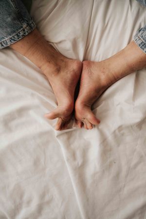 Close-up of two bare feet crossed together resting on white bed sheets, wearing blue denim jeans.