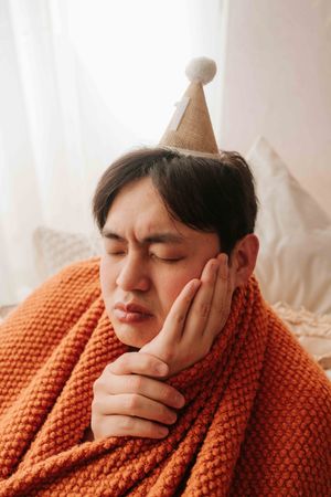 Sick Asian man wearing party hat wrapped in orange knit blanket, holding cheek in pain with eyes closed.