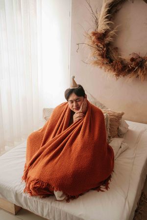 Woman wrapped in orange knit blanket sitting on bed, appearing unwell with hand on chin, autumn wreath on wall.