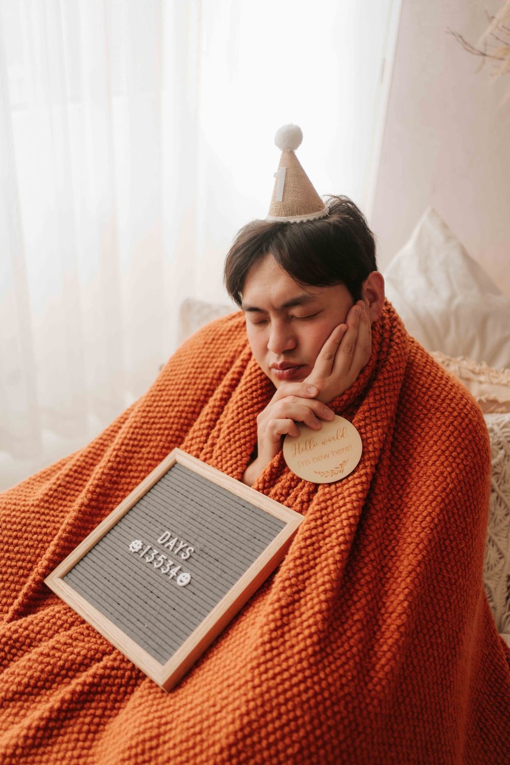 Sad man wrapped in orange blanket wearing party hat sitting on couch with letter board showing days count.