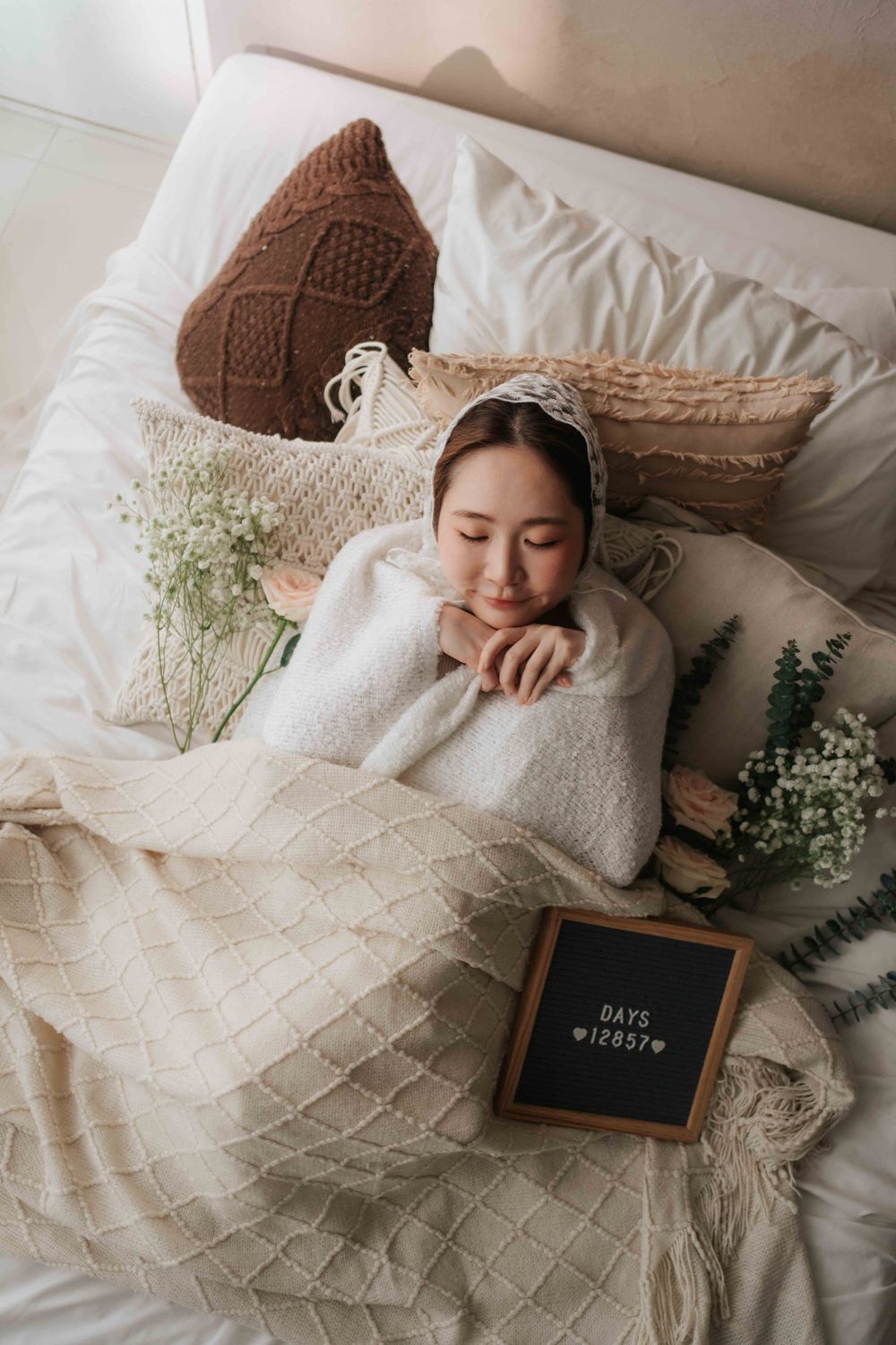 A young Asian woman lies peacefully on a white bed wrapped in a cream blanket, surrounded by flowers and a days counter sign.