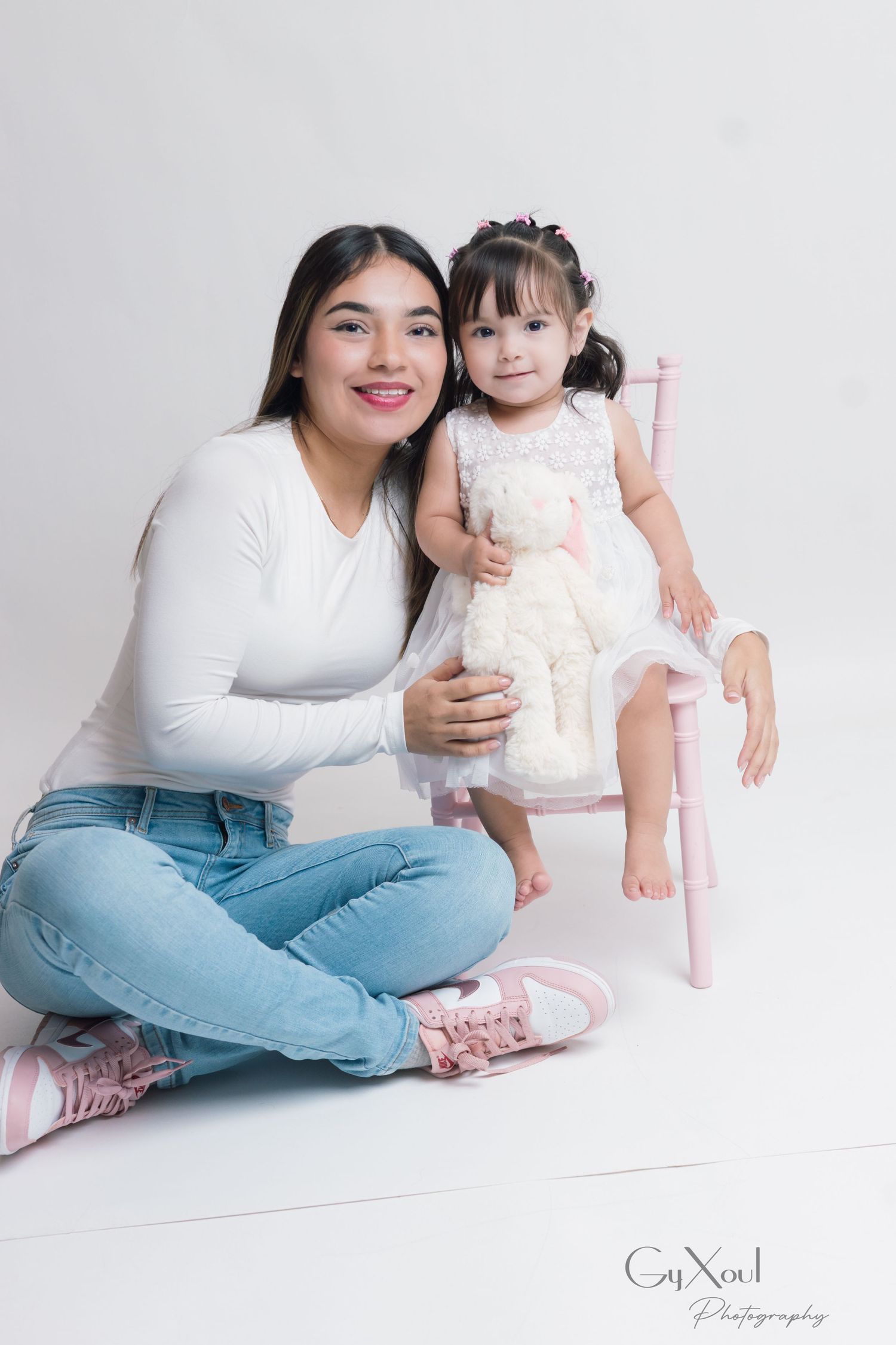 A young woman and toddler girl pose together in a white studio, the child sitting on a pink chair holding a white teddy bear.