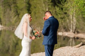 Emotional groom in navy suit wiping tears as bride in white gown holds bouquet during outdoor lakeside wedding ceremony.