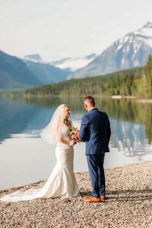 Bride and groom exchange vows on a rocky lakeshore with snow-capped mountains and a serene lake reflection behind them.
