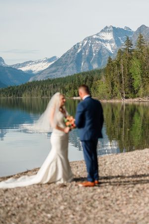 Bride and groom stand by a reflective mountain lake with snow-capped peaks and evergreen forest backdrop.