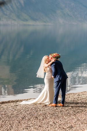 A bride and groom share a kiss by a serene mountain lake, she in a white gown with veil, he in a navy suit.