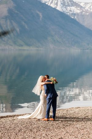 Newlywed couple shares a romantic embrace on a pebble beach with snow-capped mountains reflected in a calm alpine lake.