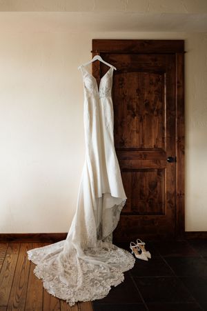Elegant lace wedding gown with long train hangs on wooden door beside white bridal heels on dark tile floor.