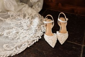 Delicate lace bridal heels with ankle straps displayed beside intricate embroidered wedding gown on dark wood floor.