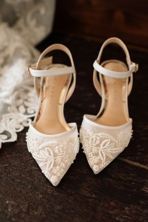 Elegant beaded white bridal heels with ankle straps and floral pearl embellishments on dark wood surface.