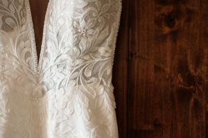 Close-up detail of elegant ivory lace wedding dress with deep V-neckline hanging against rustic wood paneled wall.