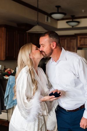 Bride in white feathered robe and groom in white shirt share a kiss while holding a ring box in kitchen.