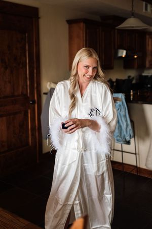 Smiling bride in monogrammed white feather-trimmed satin robe holding phone while getting ready indoors.