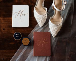 Wedding flat lay featuring lace heels, his and her vow books, rings, and bridal veil.