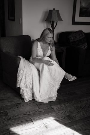 Bride in lace wedding gown sits in armchair adjusting her shoe in a black and white portrait.