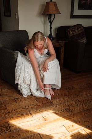 Bride in white wedding dress putting on heels while seated on gray chair on hardwood floor.