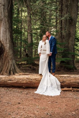Bride and groom pose among tall trees in a forest, bride wearing a white gown with lace train and fur jacket.